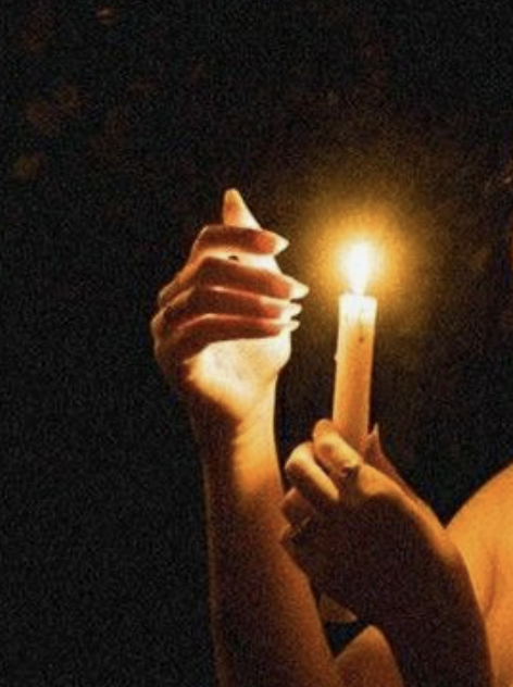 A person holding a lit candle in a dark setting, with their hand near their face.