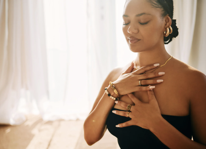 A woman with closed eyes and a serene expression, wearing a strapless black top, gold jewelry, and bracelets, standing indoors near a window with curtains.