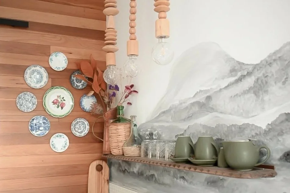 Wall with a mural of mountainous landscape, decorated with green ceramics and glass bottles on a wooden shelf, with a cluster of decorative plates on a wooden wall, and dried flowers in a basket.