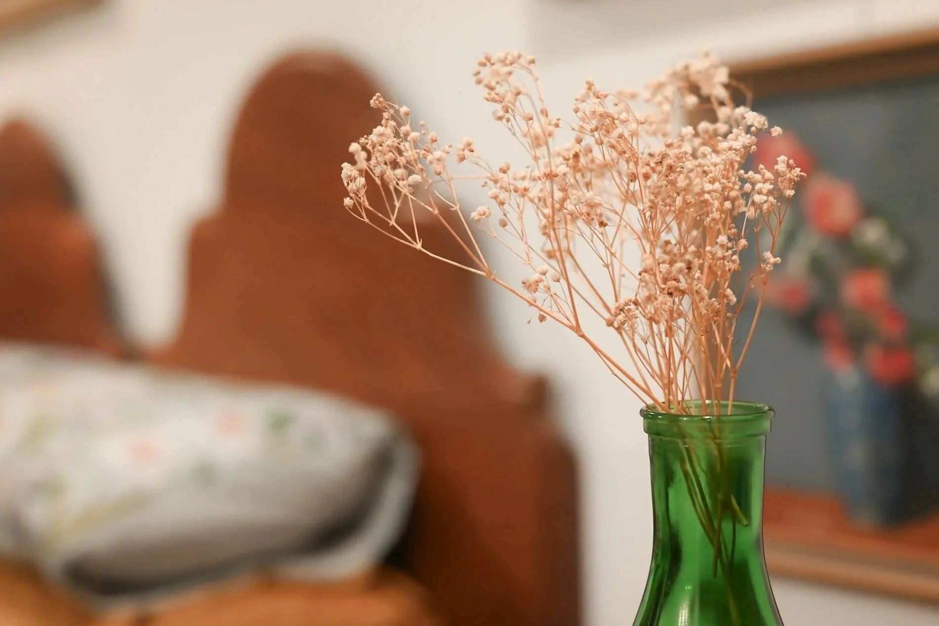Dried flowers in a green glass vase on a table, blurred background of floral paintings on the wall and bedhead.