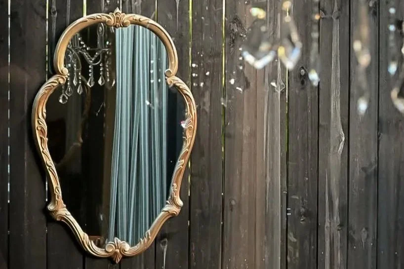 Decorative mirror with a gold ornate frame hung on a dark wooden wall, partially reflecting a blue curtain and a chandelier with crystal accents.