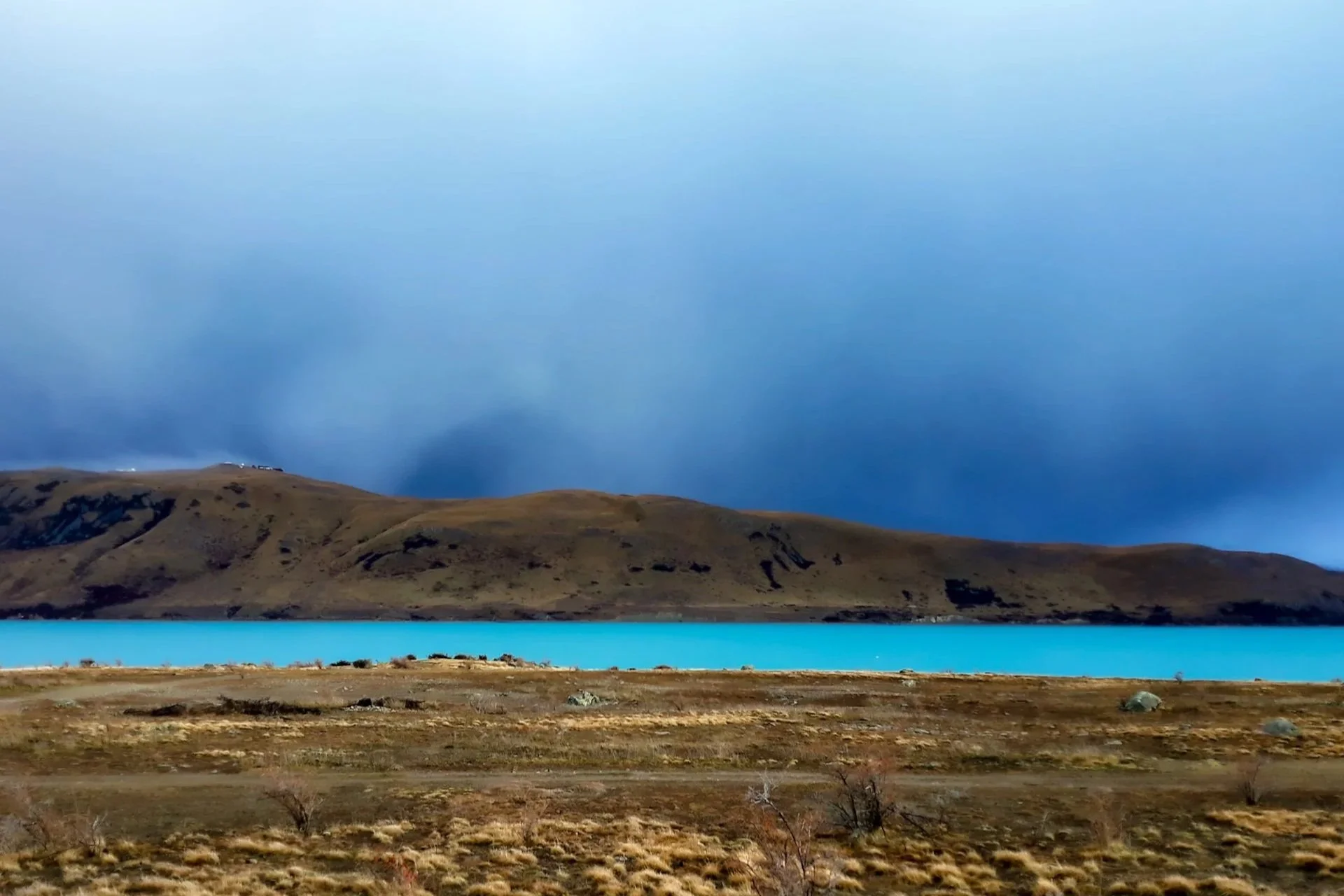 A landscape featuring a calm turquoise lake, rolling brown hills, and a cloudy blue sky.