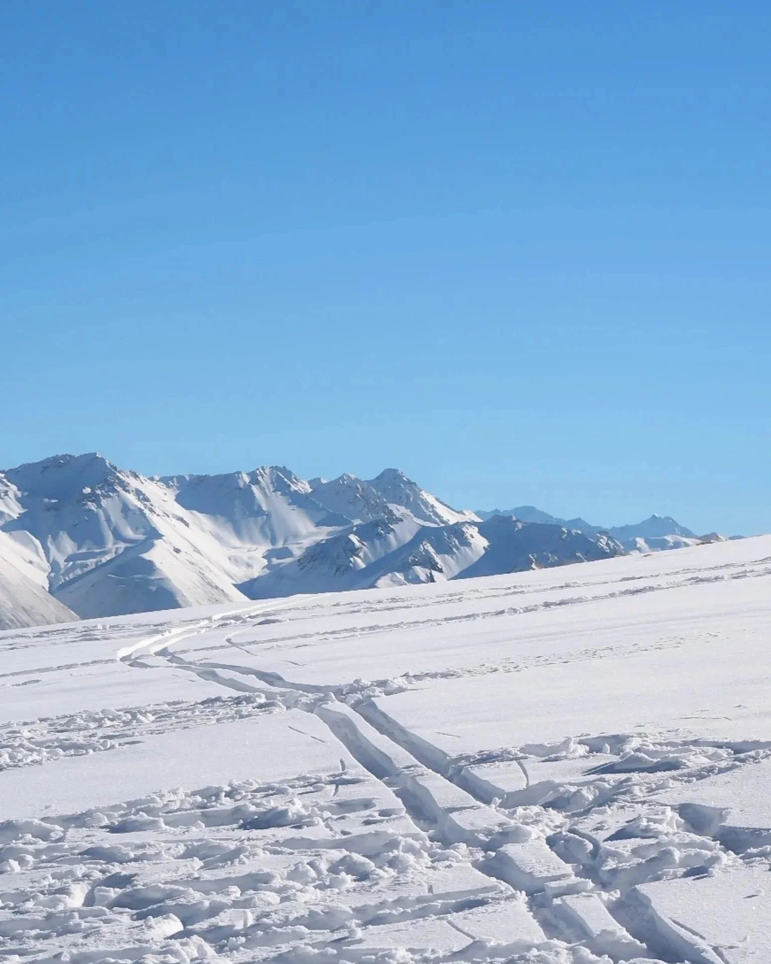 Snow-covered mountain landscape under a clear blue sky, with ski tracks in the foreground.