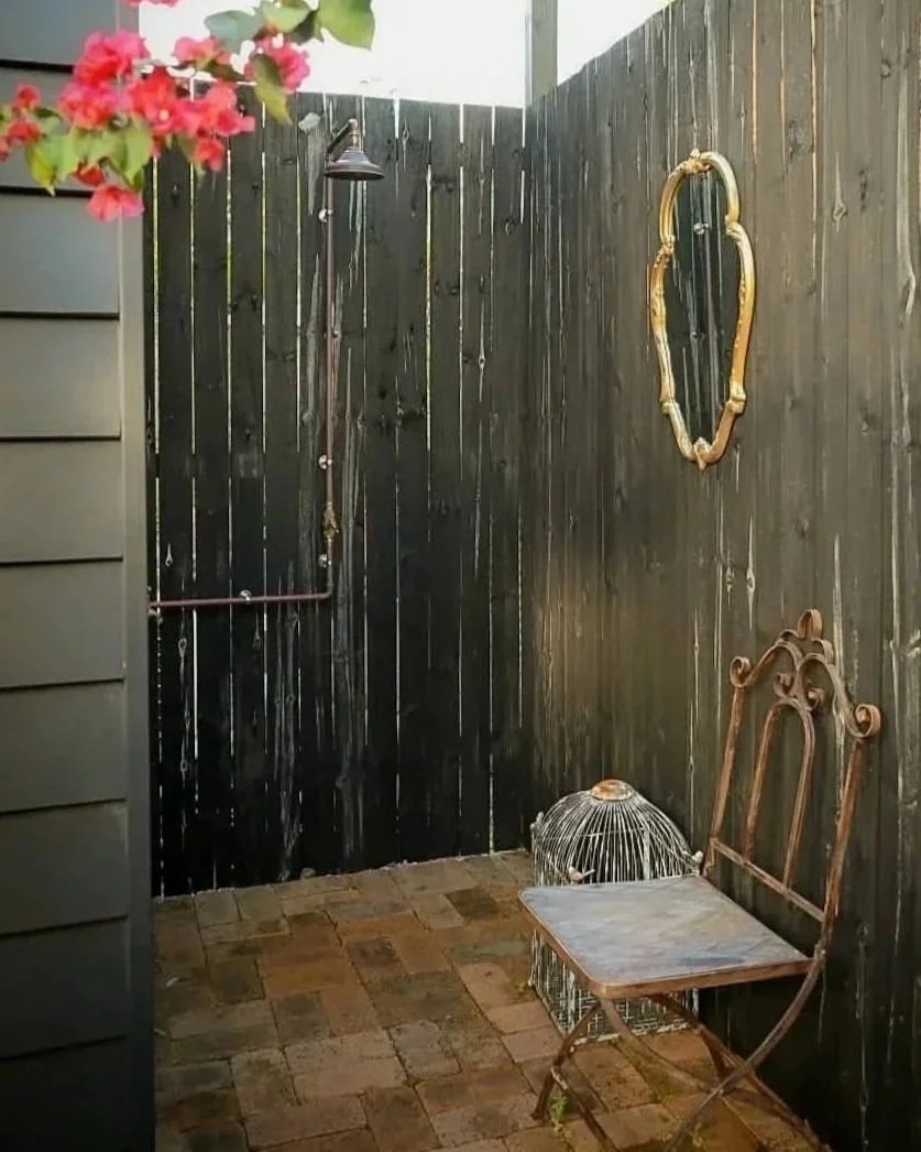 An outdoor shower area with black wooden walls, a brass mirror on the wall, a vintage metal chair with a stone seat, and a birdcage on the ground. Pink flowers are visible in the top left corner.