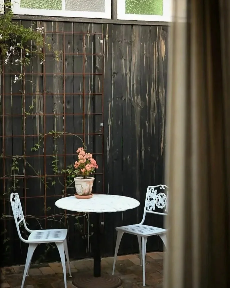 A small outdoor patio scene with a white round table, two white decorative chairs, and a potted pink flowering plant on the table, enclosed by a black wooden fence with plants climbing on a trellis, seen through a partially opened curtain.
