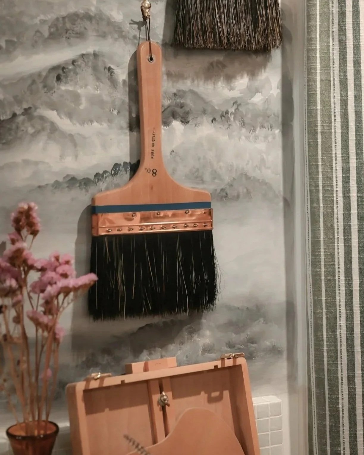 A large black and brown cleaning brush hanging on a textured gray wall, with a small brass bell attached to its handle, and a box with pink and green flowers in the foreground.