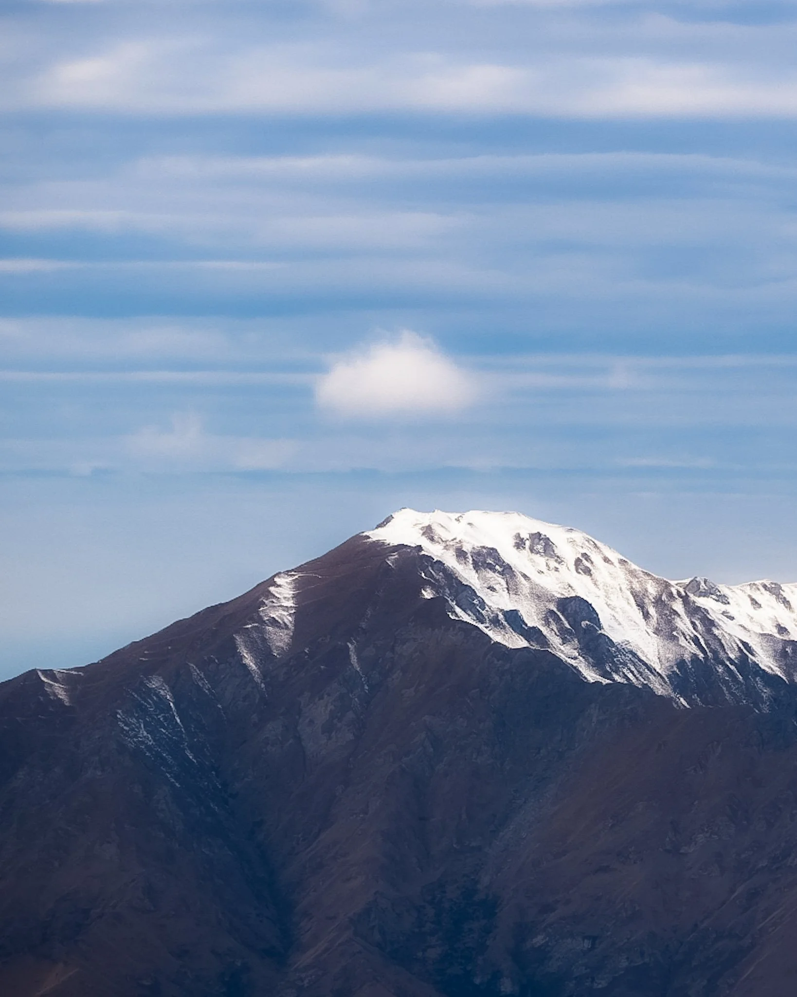 Snow-capped mountain with brown ridges under a blue sky with wispy clouds.