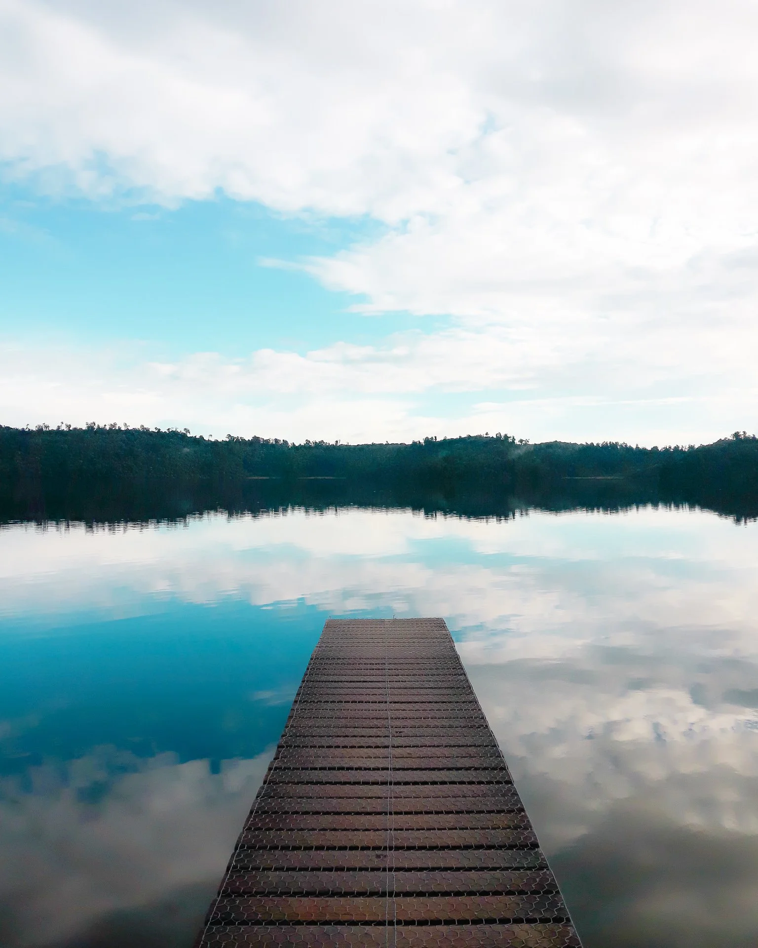 A view of a dock extending into a calm lake or river, with the water reflecting the cloudy sky and surrounding trees.