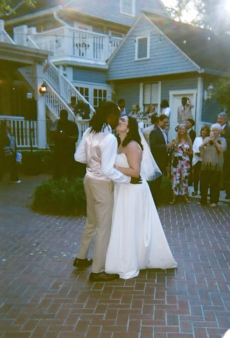 A bride and groom dance closely together at their wedding reception outside a blue house, with guests watching and a house with stairs and a deck in the background.