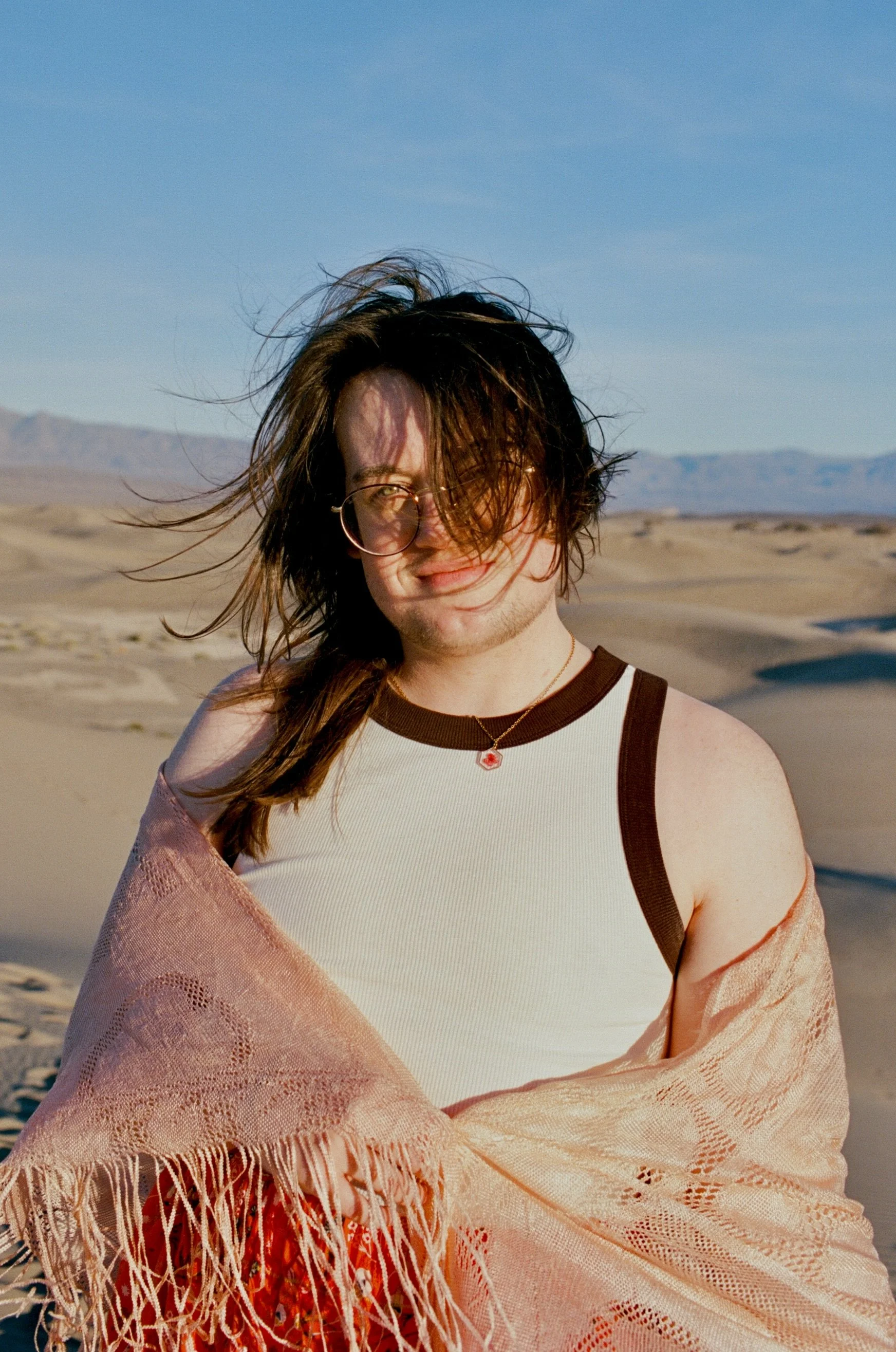 A person with long, windswept hair standing in a desert landscape with sand dunes and mountains in the background, wearing glasses, a sleeveless shirt, and a shawl.