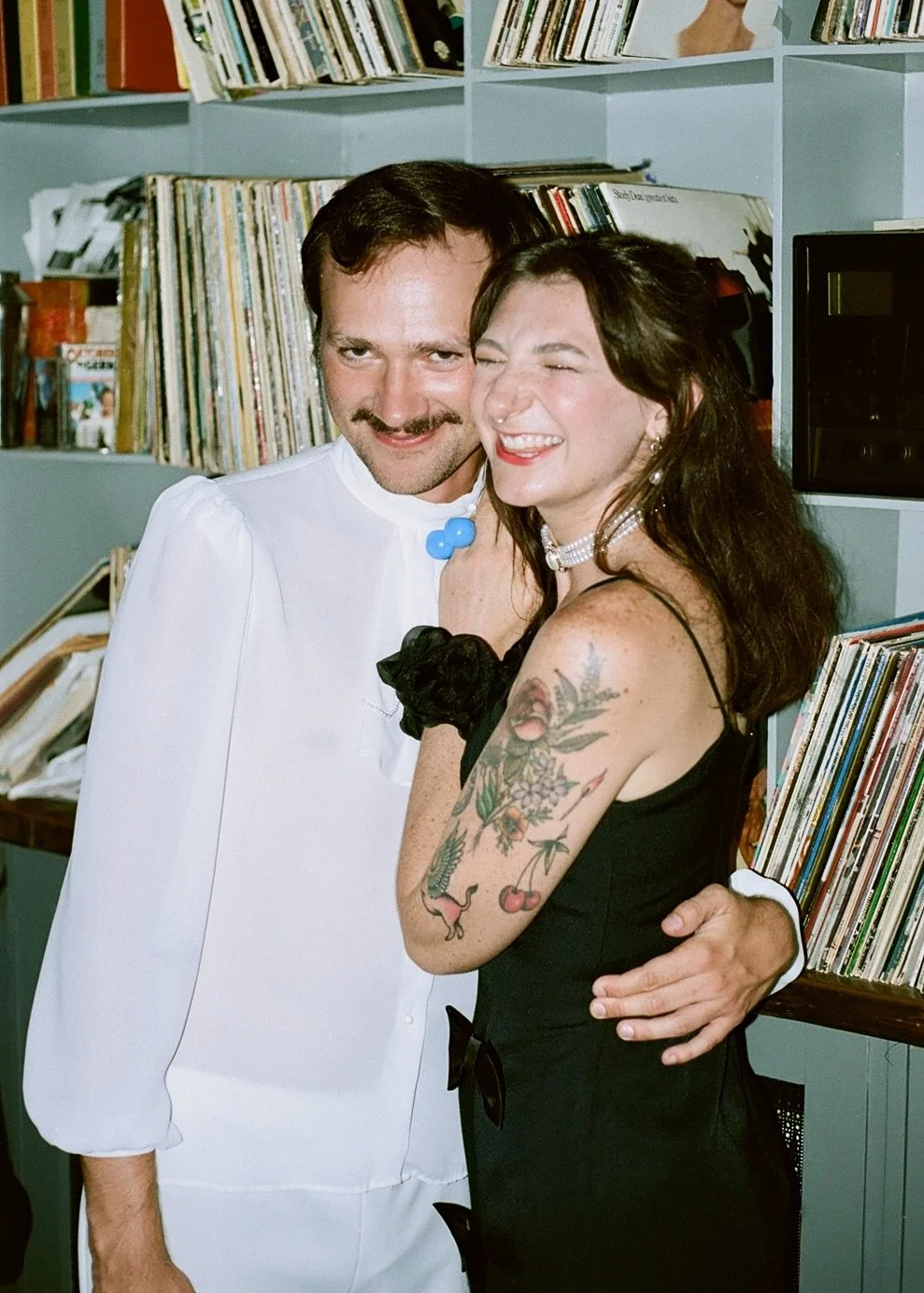 A man in a white outfit and a woman in a black dress are smiling and hugging each other in front of a bookshelf filled with records at a record store or library. The woman has tattoos on her arm, dark hair, red lipstick, and is wearing jewelry.