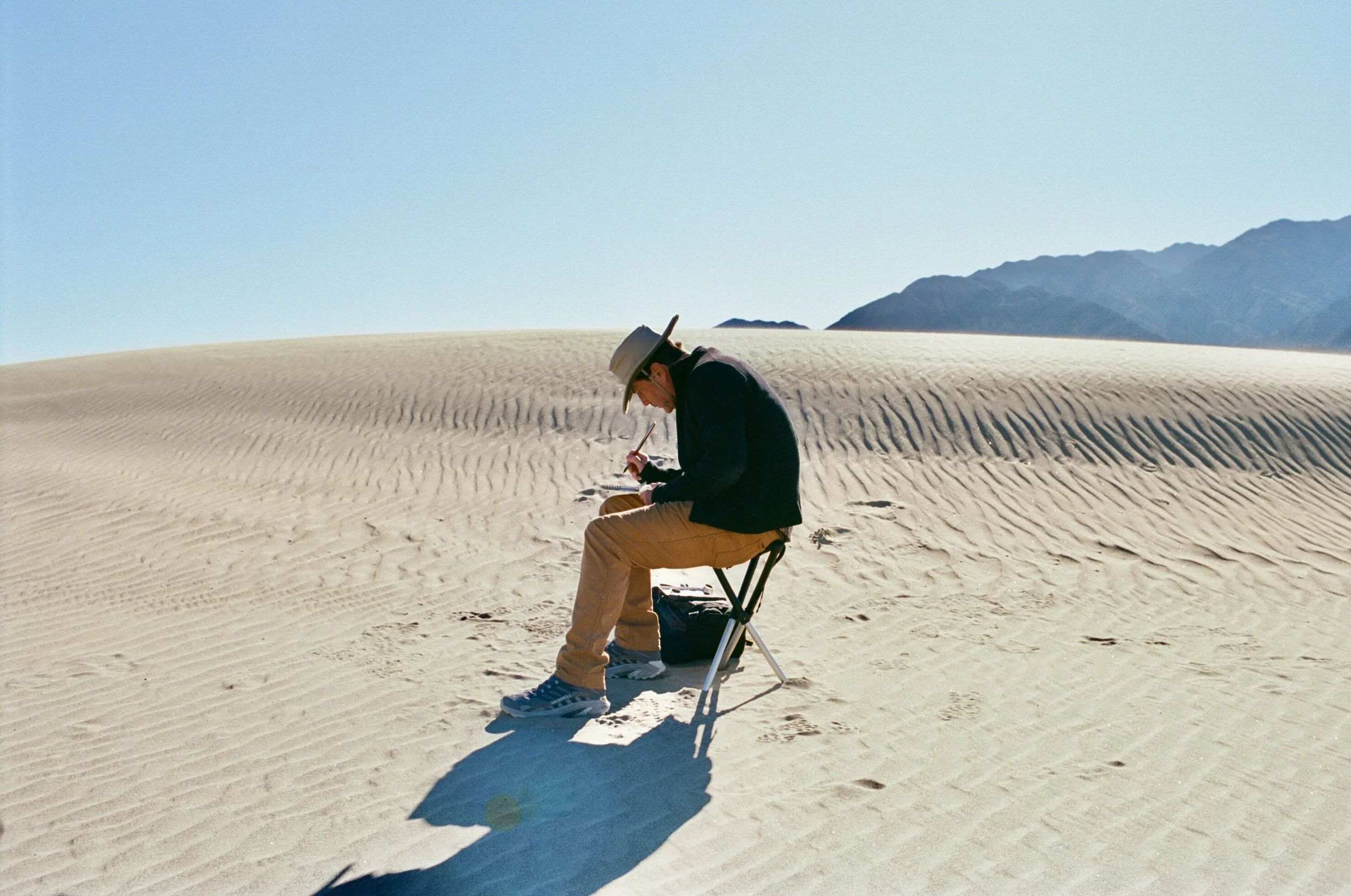 A person wearing a wide-brimmed hat, black jacket, tan pants, and sneakers sitting on a folding chair in a desert, writing or drawing on a notepad with mountains in the background.