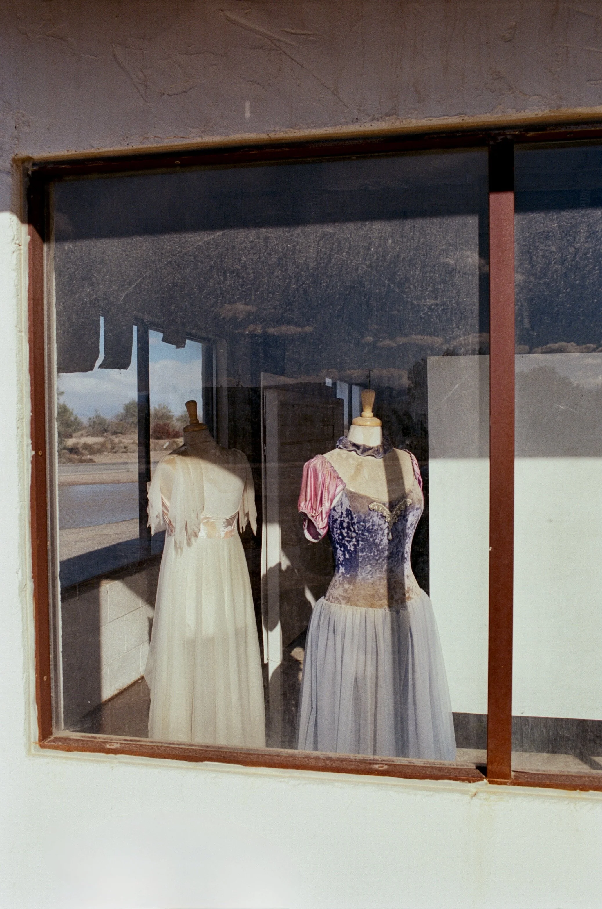 Two vintage dresses displayed on mannequins in a dusty storefront window, with a landscape and river seen outside.