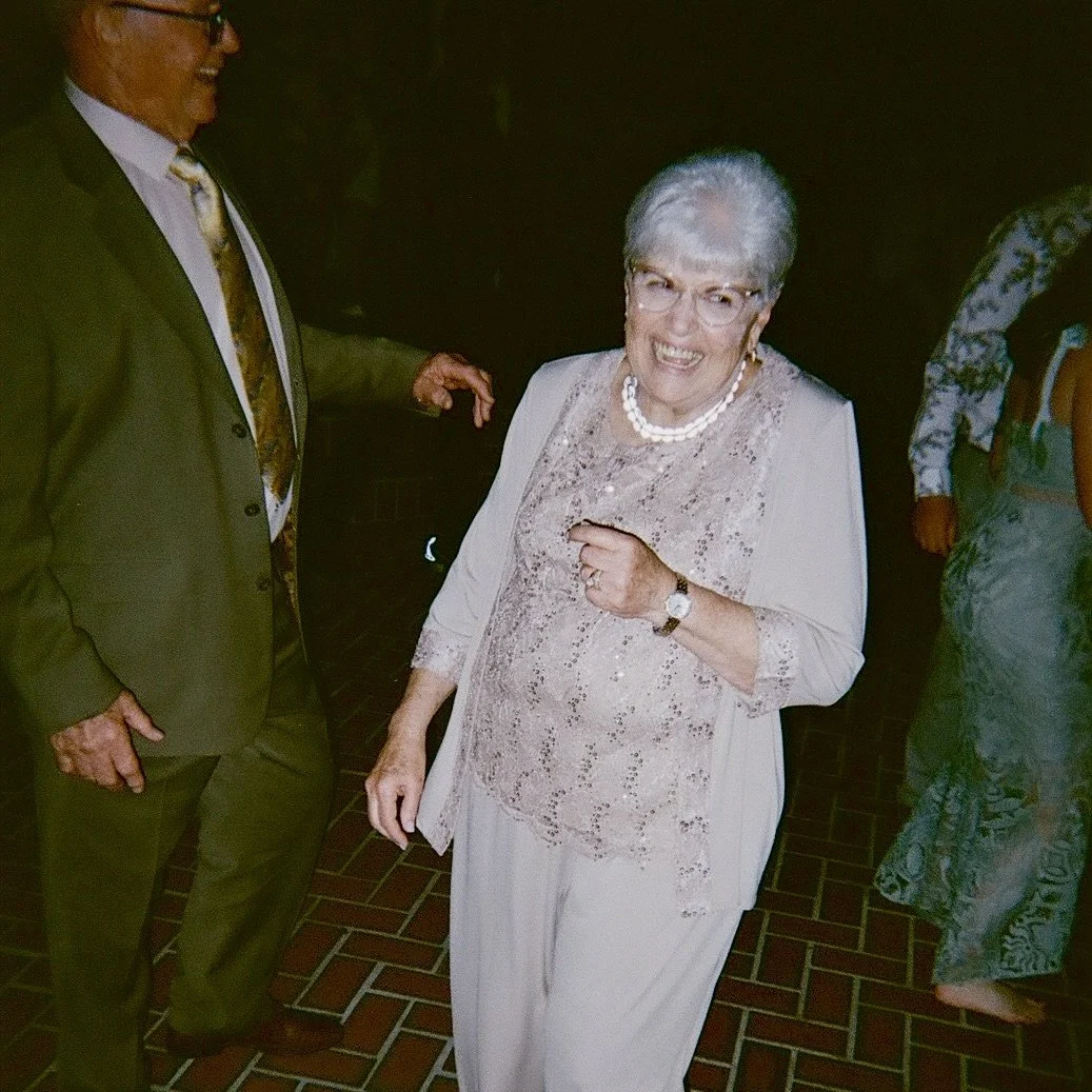 An elderly woman with gray hair and glasses, smiling and pointing at the camera, wearing a light-colored dress with lace details, pearl necklace, watch, and rings, standing on a brick floor at a social event.