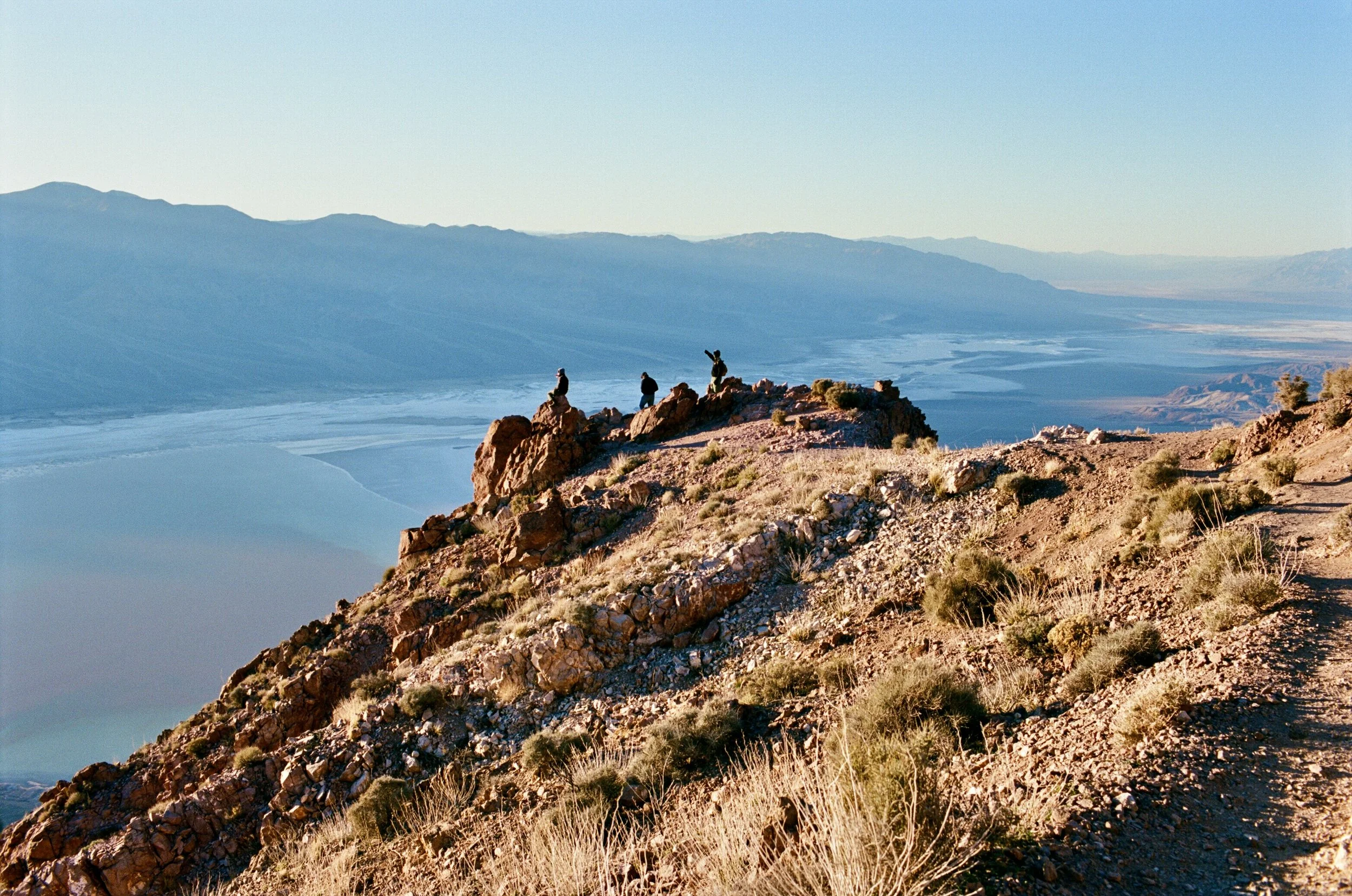 Three people sitting on rocky outcrop overlooking a desert landscape with mountains in the background during sunset.