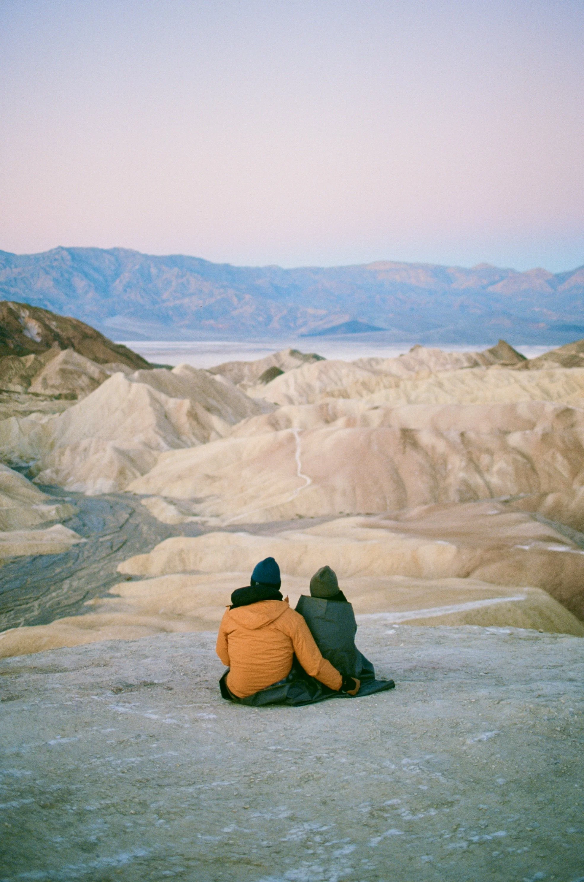 Two people sitting on a rocky foreground, overlooking a desert landscape with mountains in the background at dusk.