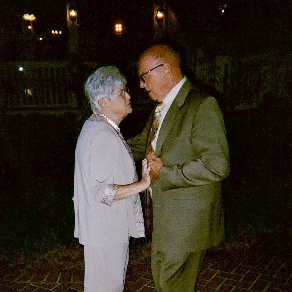 An elderly woman and a middle-aged man are dancing closely, facing each other, in a dimly lit room.
