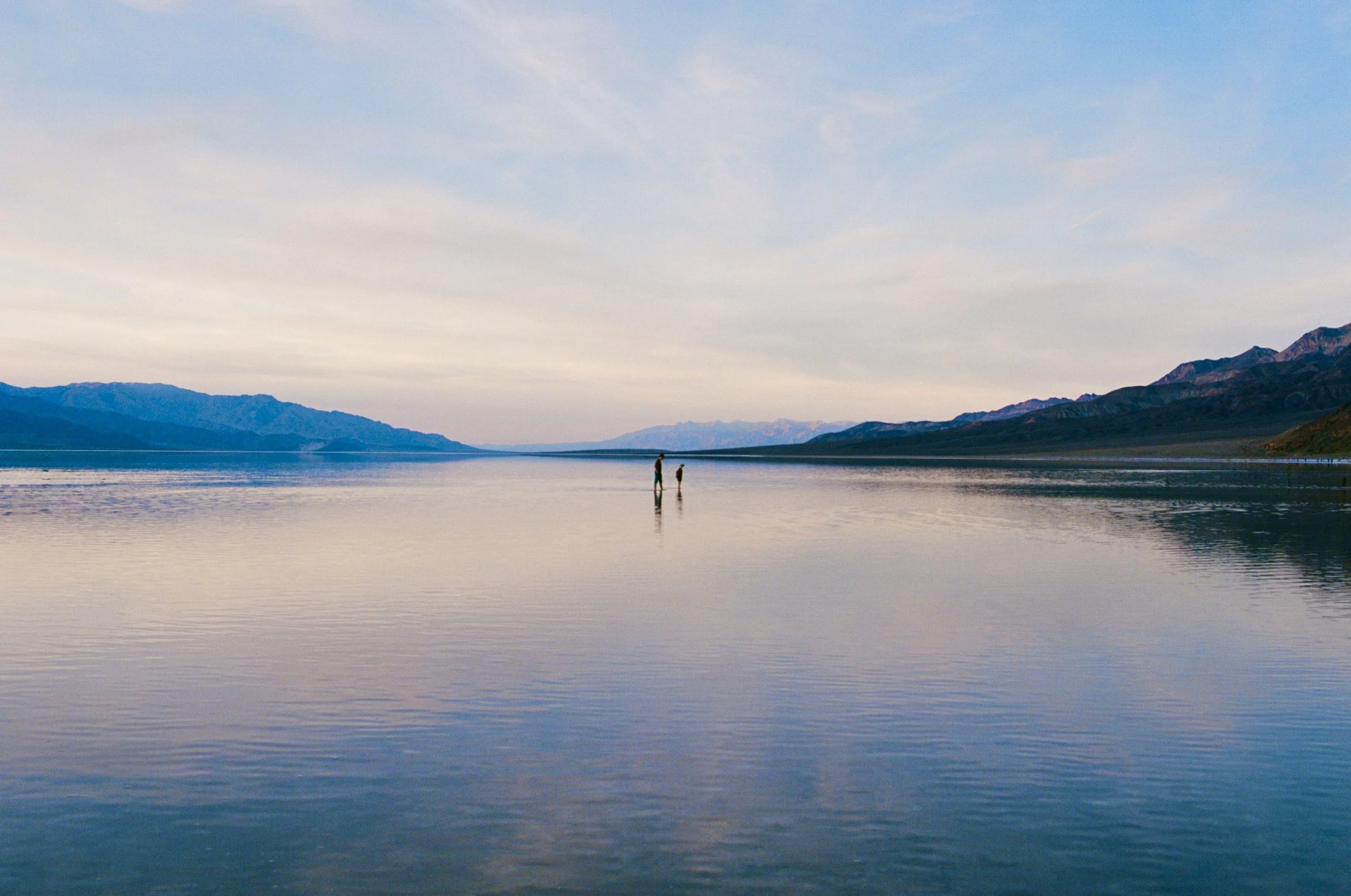 Two people stand in shallow water at a vast lake, with mountains in the background and a sky filled with soft clouds.