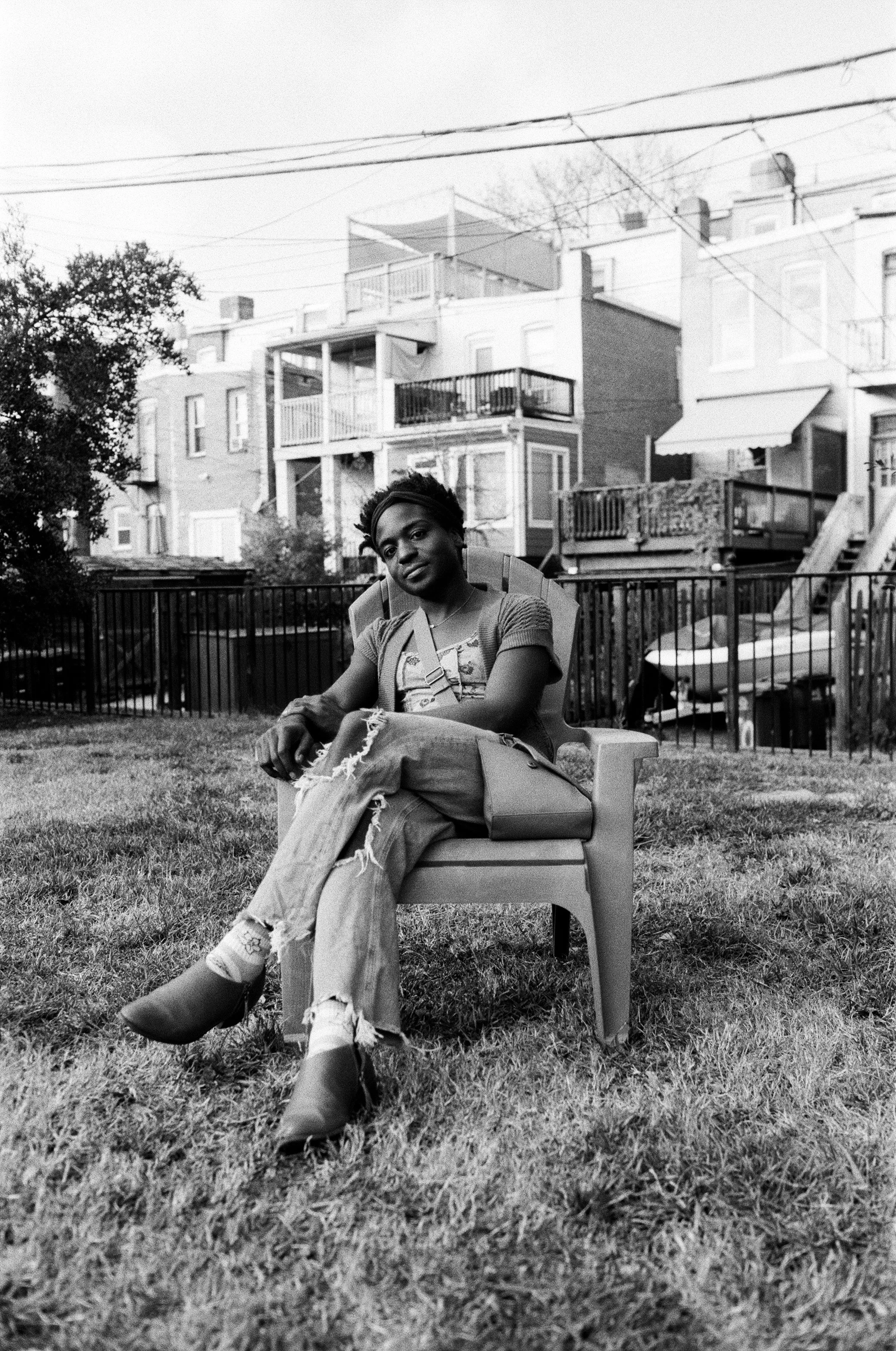 A woman sitting on a lawn chair in a backyard, with residential buildings and balconies in the background.