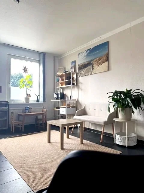 Gemütliches Wohnzimmer mit weißem Sofa, Couchtisch, Bücherregal, Topfpflanzen, Wanddekoration und Fenster mit Blick nach draußen.