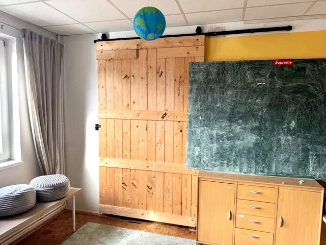 Klassenzimmer mit grüner Tafel, einem Holzschrank, einem Globus an der Decke, einem Fenster mit Vorhängen, zwei gestreiften Kissen auf einer Bank und einem Regal.