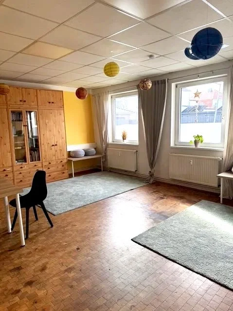 Geräumiges Zimmer mit Holzschrank, zwei großen Fenstern mit Vorhängen, zwei Teppichen, einem Stuhl, einem kleinen Tisch und Dekorationen an der Decke, darunter bunte Laternen.
