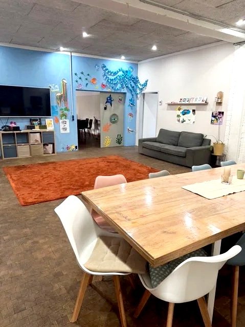 Gemütlicher Gemeinschaftsraum mit Fernseher, Kinderkunst an der Wand, graues Sofa, Holz-Tisch mit Stühlen, roter Teppich, Dekorationen für Kinder im Hintergrund.