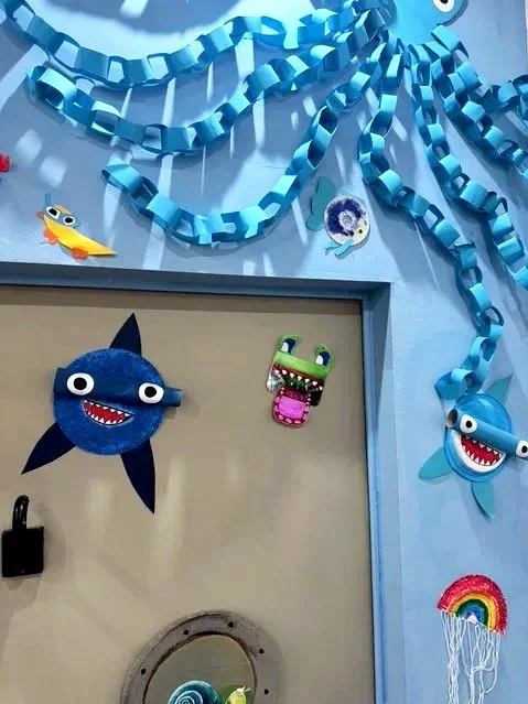 Bunte, bemalte Pappfiguren und Drahtkunstwerke in Form von Meereswesen wie ein Hai, ein Delfin, eine Schildkröte, ein Rochen und andere Meerestiere, die an einer blauen Wand und einem Türrahmen angebracht sind.