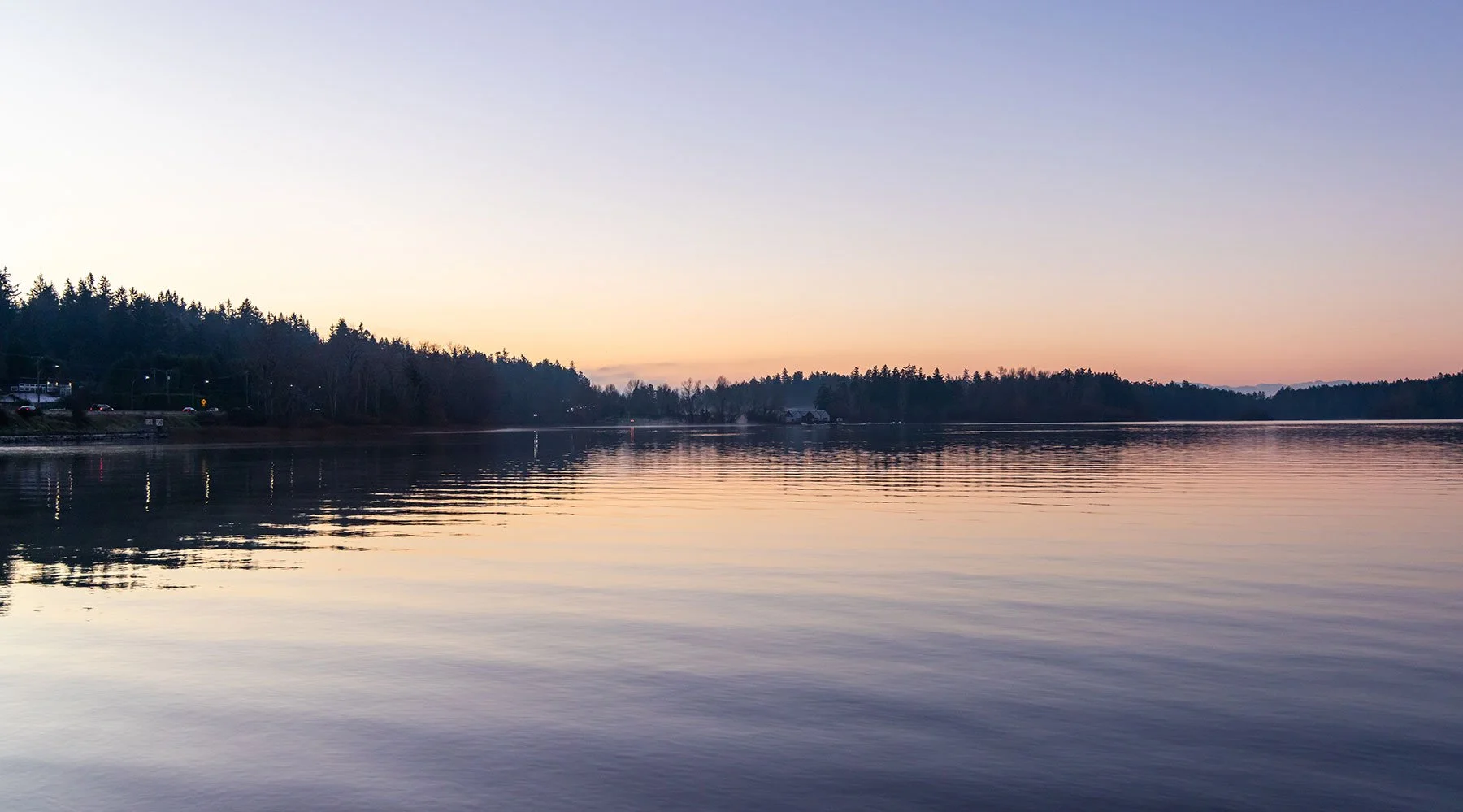 Ein ruhiger See bei Sonnenuntergang, umgeben von Bäumen und einem kleinen Haus am Horizont.