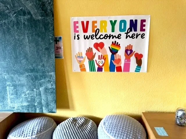 Ein Schild mit bunten Buchstaben, das sagt 'JEDER ist hier willkommen' und zeigt bunte Hände mit Regenbogen- und Herzmotiven, an einer gelben Wand im Innenraum.