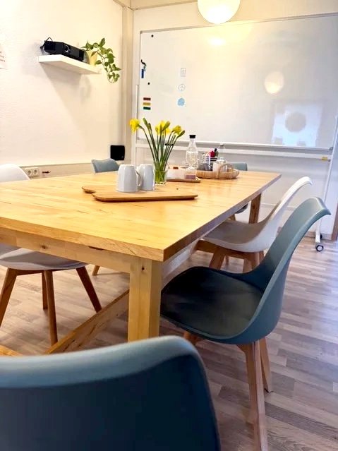 Ein Konferenztisch mit acht Stühlen, eine Vase mit gelben Tulpen, Wasserflasche, Kaffeetassen, und ein Whiteboard im Hintergrund in einem modernen Raum.