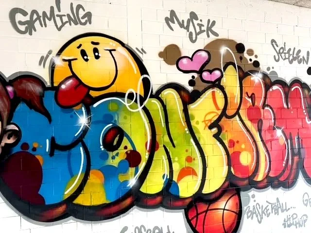 Graffiti an einer Wand mit einem lachenden Smiley, Händen, Herzsymbolen, und sportlichen Elementen, darunter ein Basketball.