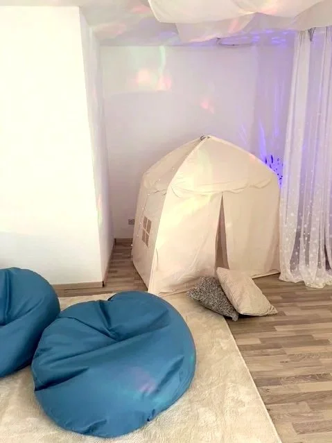 Ein Indoor-Peak-Zelt auf einem Holzboden, umgeben von blauen Sitzsack-Stühlen und einem Vorhang.