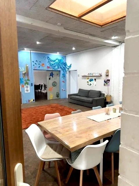 Ein gemütlicher Raum mit Esstisch und Stühlen, Wohnzimmer mit Sofa und bibliotheksähnlichen Regalen, bunte Wanddekorationen und eine Fensterdecke. Verschiedene Möbel und Dekorationsgegenstände sind sichtbar.