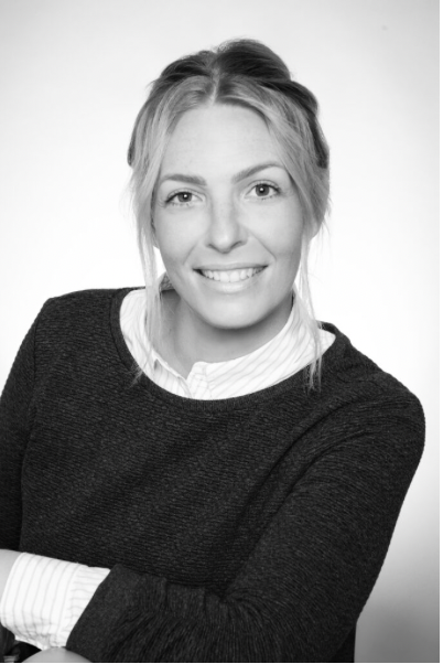Schwarz-weiß-Fotografie eines lächelnden Frauenporträts, mit lockigem Haar, in casual Kleidung, vor hellem Hintergrund.