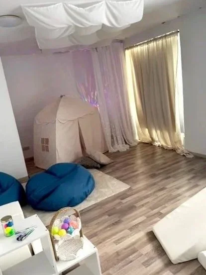Ein Kinderzimmer mit Zelt, zwei blaue Sitzsacke, einem Spielzeugkorb mit bunten Bällen, weißen Möbeln und Vorhängen vor einem Fenster.