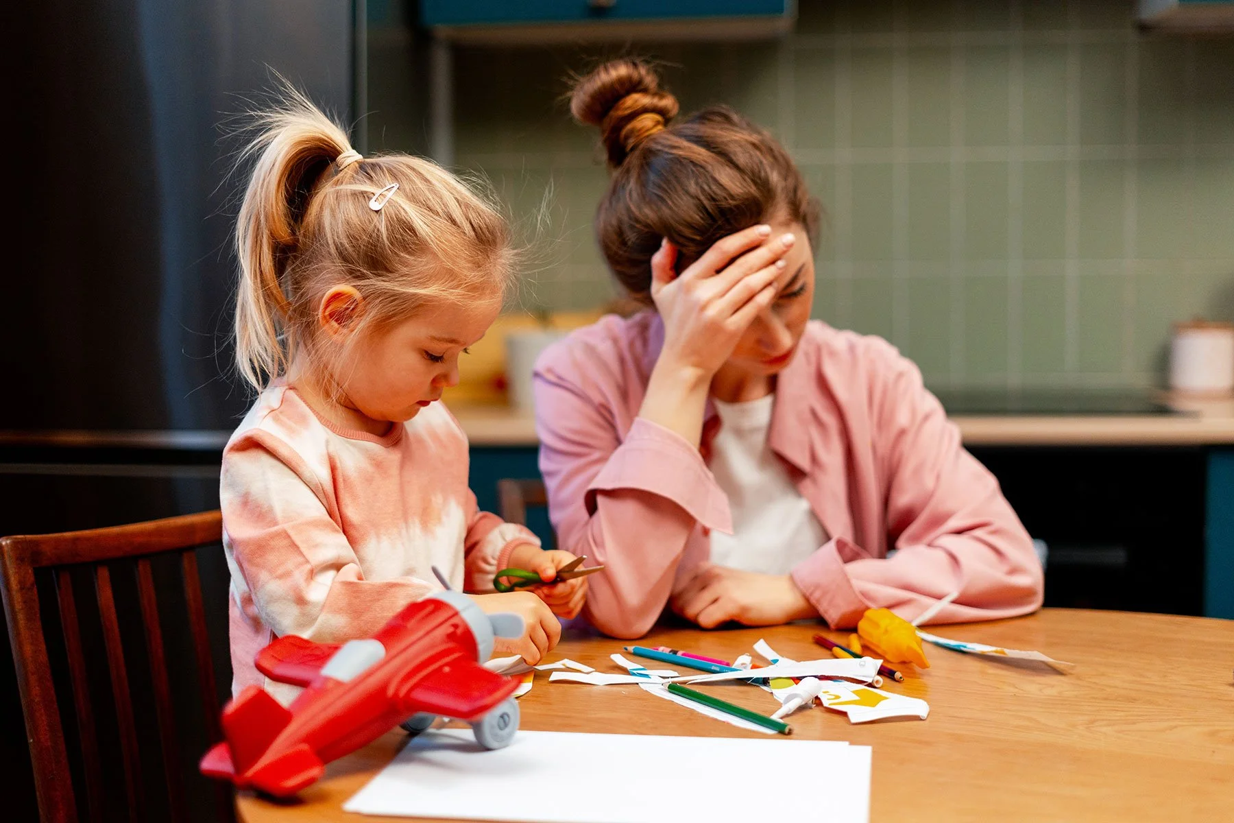 Zwei junge Mädchen sitzen an einem Holztisch, eines sieht verärgert aus, während sie an Bastelarbeiten mit Papier, Schere und Klebstoff arbeiten.