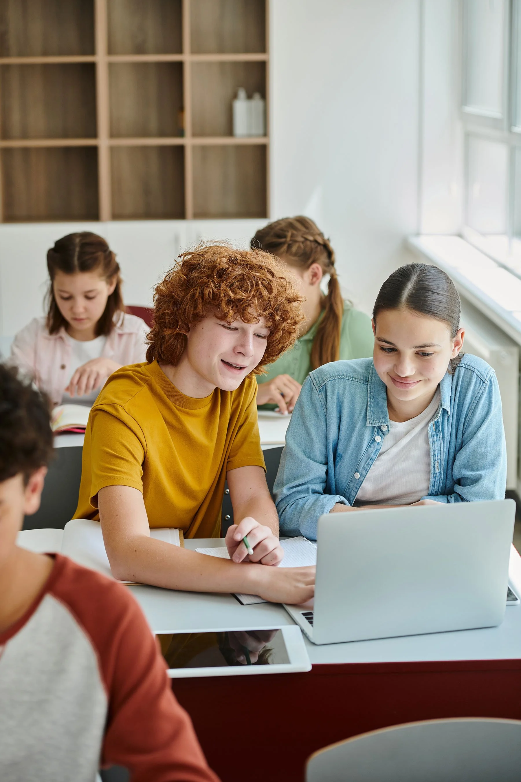 Junge Mädchen und Jungen sitzen um einen Tisch in einem Klassenraum, schauen auf einen Laptop, während sie gemeinsam lernen oder arbeiten.