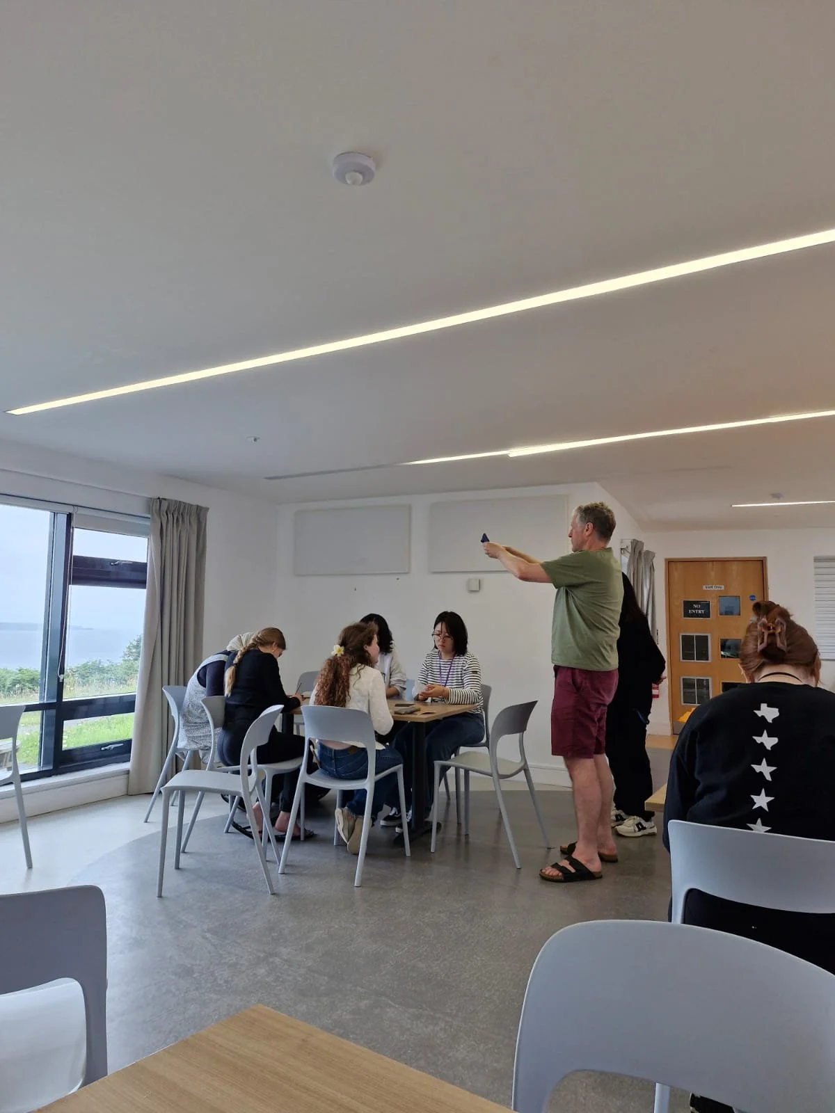 A group of people seated around a table in a room with large windows, while a man stands and points with a pen or pencil. The room has white walls, beige curtains, and a view of greenery outside.