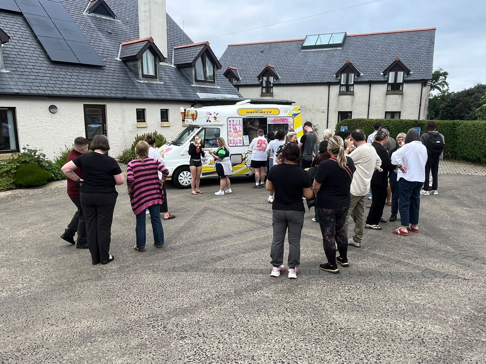 A group of people gathered outside a building, with some standing near a colorful ice cream truck decorated with cartoon characters. The background shows a house with a dark roof and skyline overcast sky.