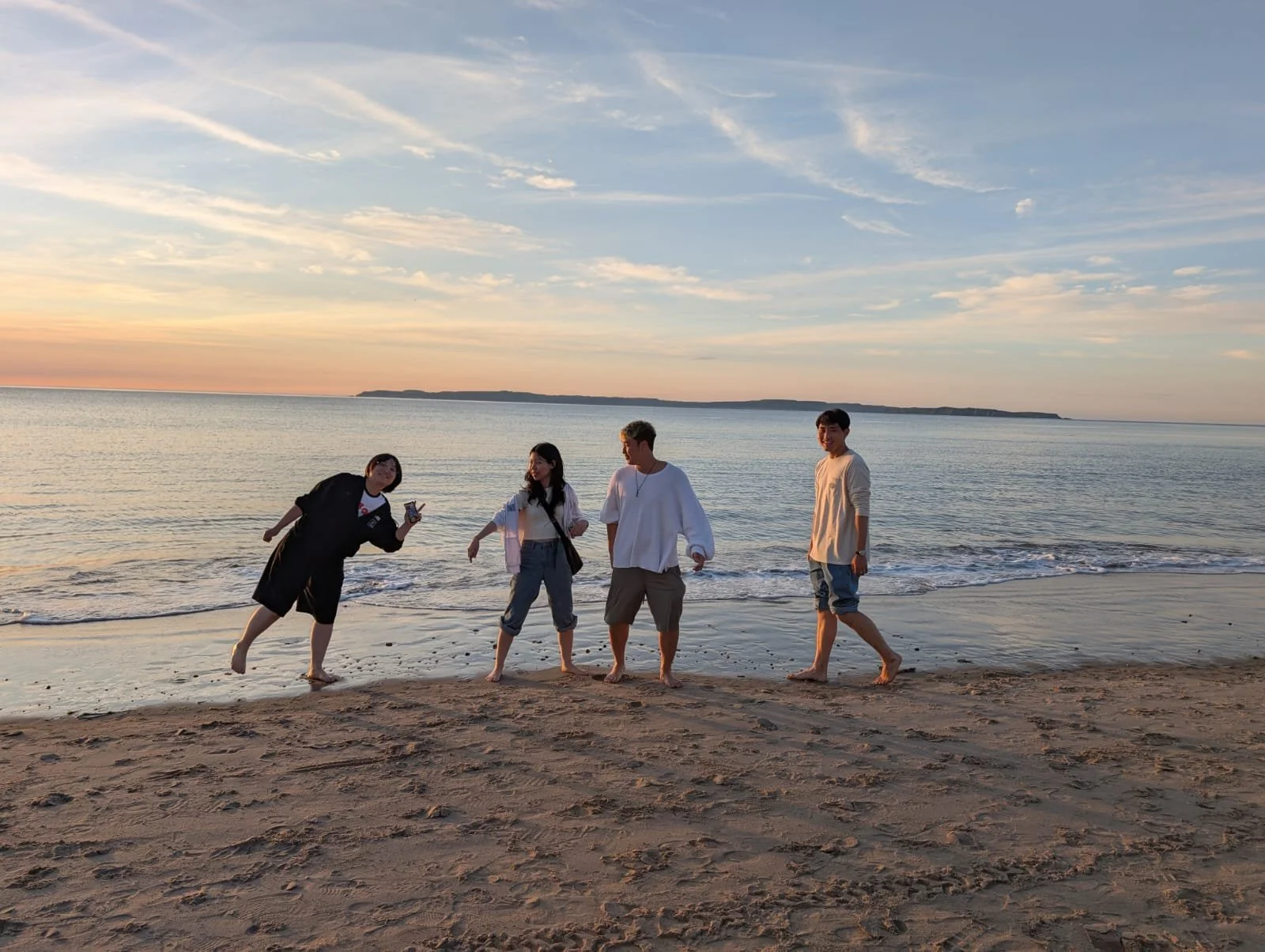 Four young people standing on a sandy beach at sunset, with calm ocean waves behind them and a distant landmass on the horizon.