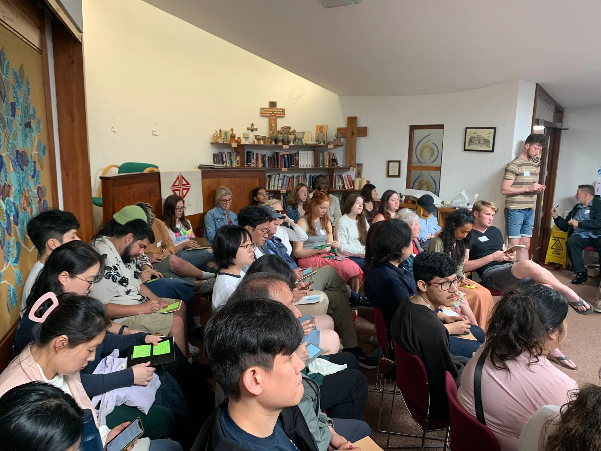 A group of people seated and reading or taking notes in a room with religious decorations, including crosses, bookshelves, and framed artworks.
