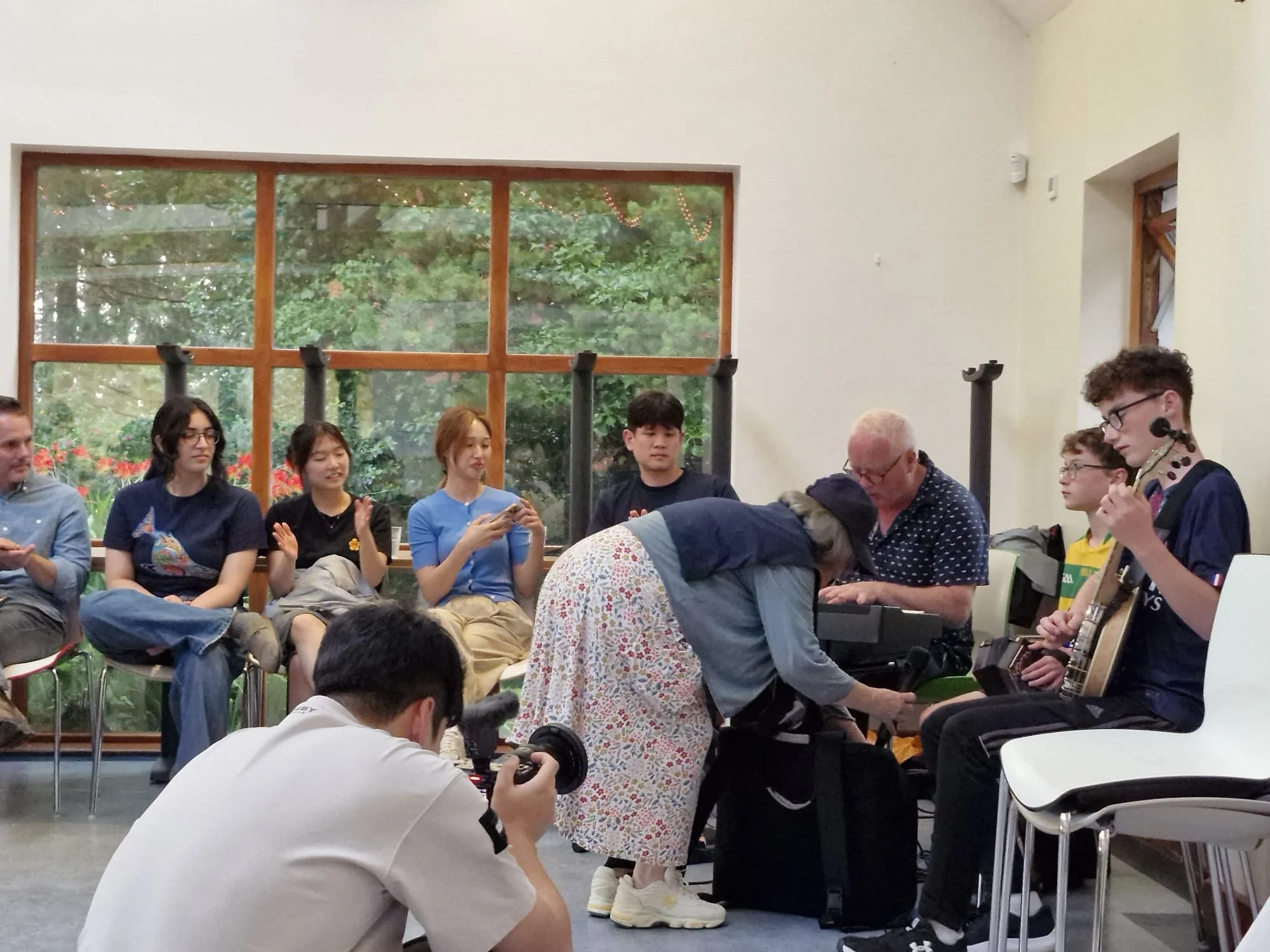 People sitting and preparing for a musical performance indoors, with a large window showing trees outside.