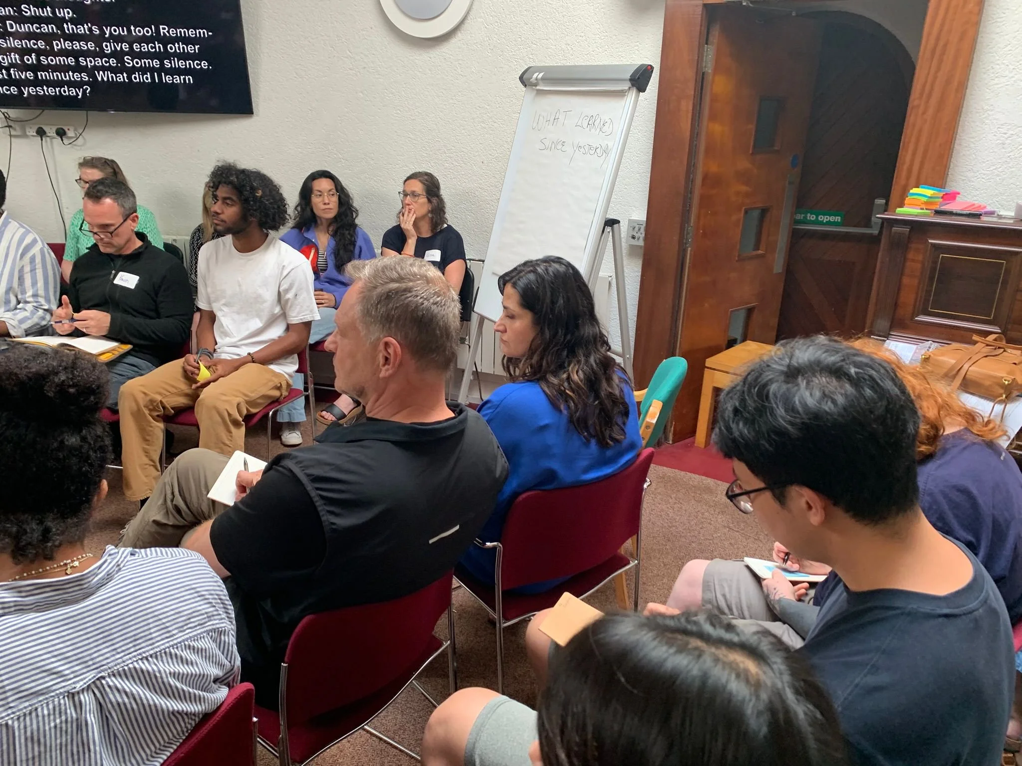 People sitting in a circle in a room, participating in a discussion or workshop. A whiteboard with handwritten notes is visible, and a large screen on the wall displays partially visible text.