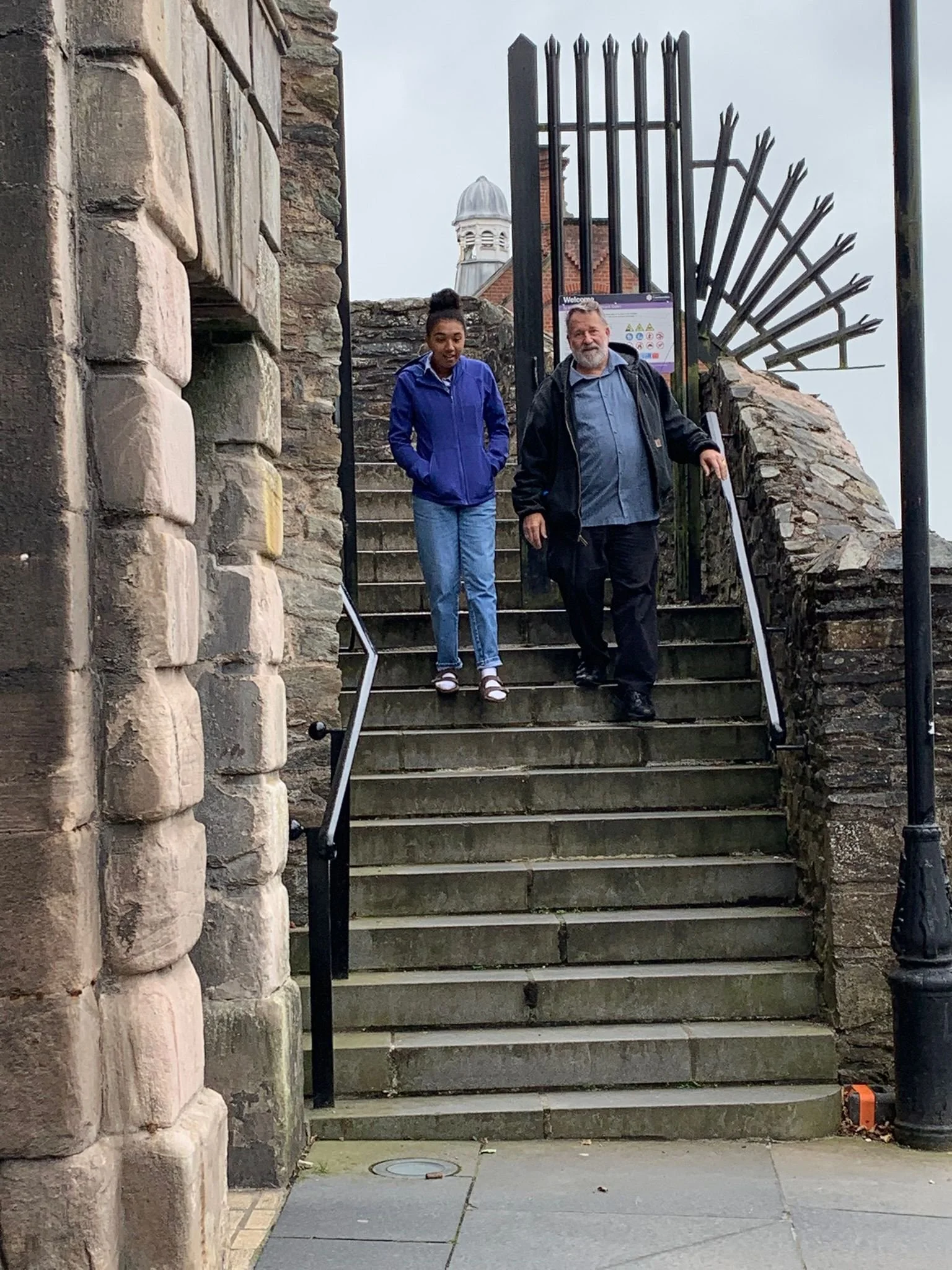 Two people, a young woman and an older man, walking down stone steps next to a brick wall and black iron gate on an overcast day.