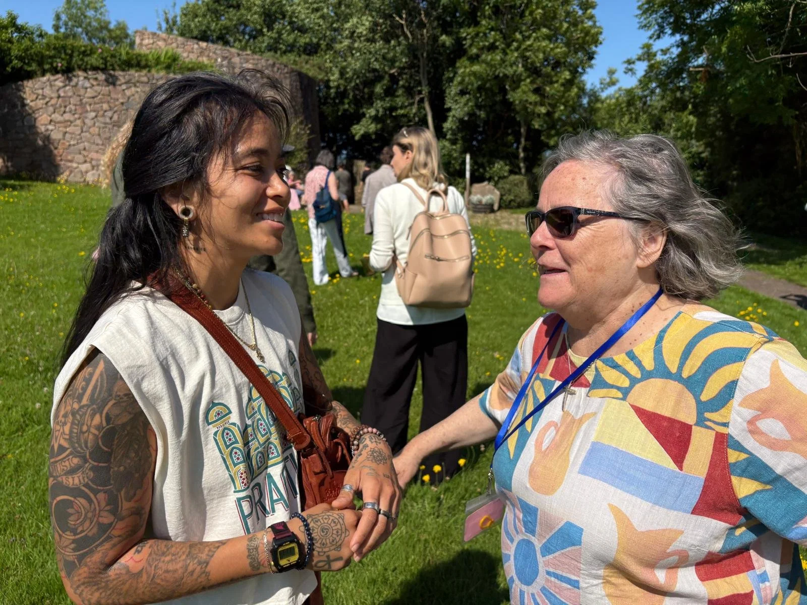 Two women are smiling and shaking hands outside the Croi, with other people and green trees in the background.
