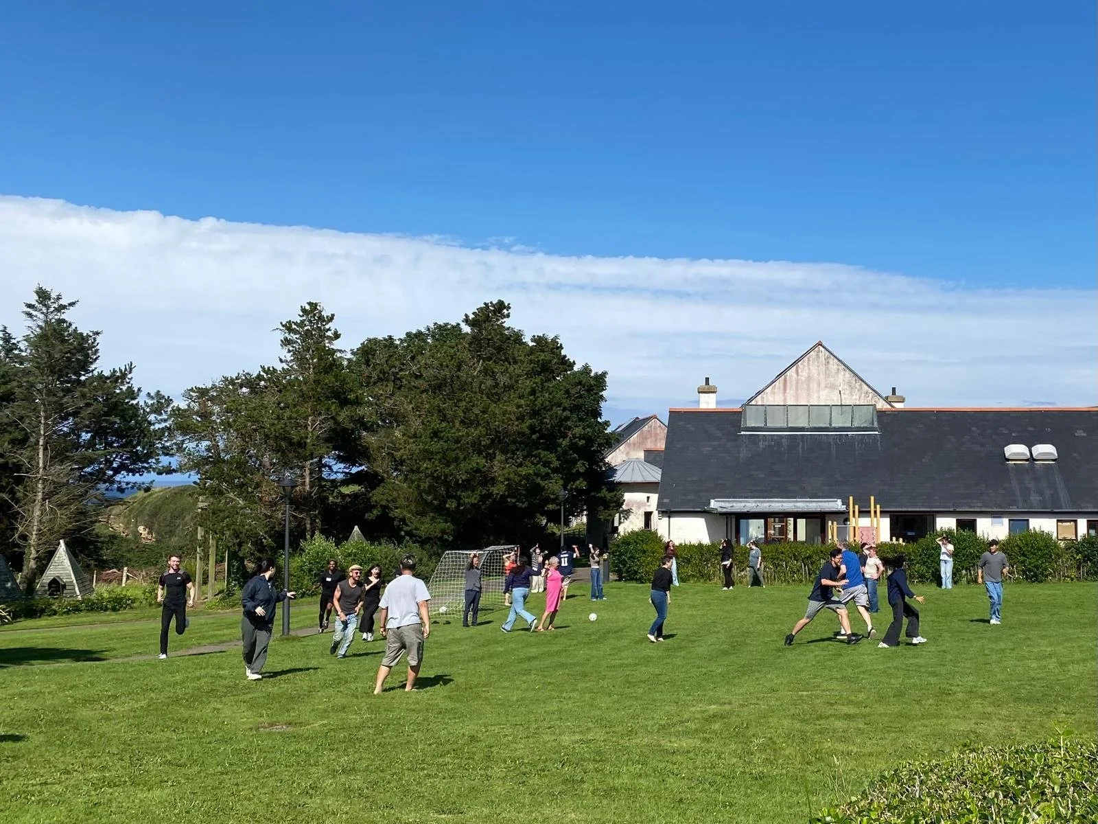 People playing soccer on a grassy field near a house with a black roof, surrounded by trees and blue sky with some clouds.