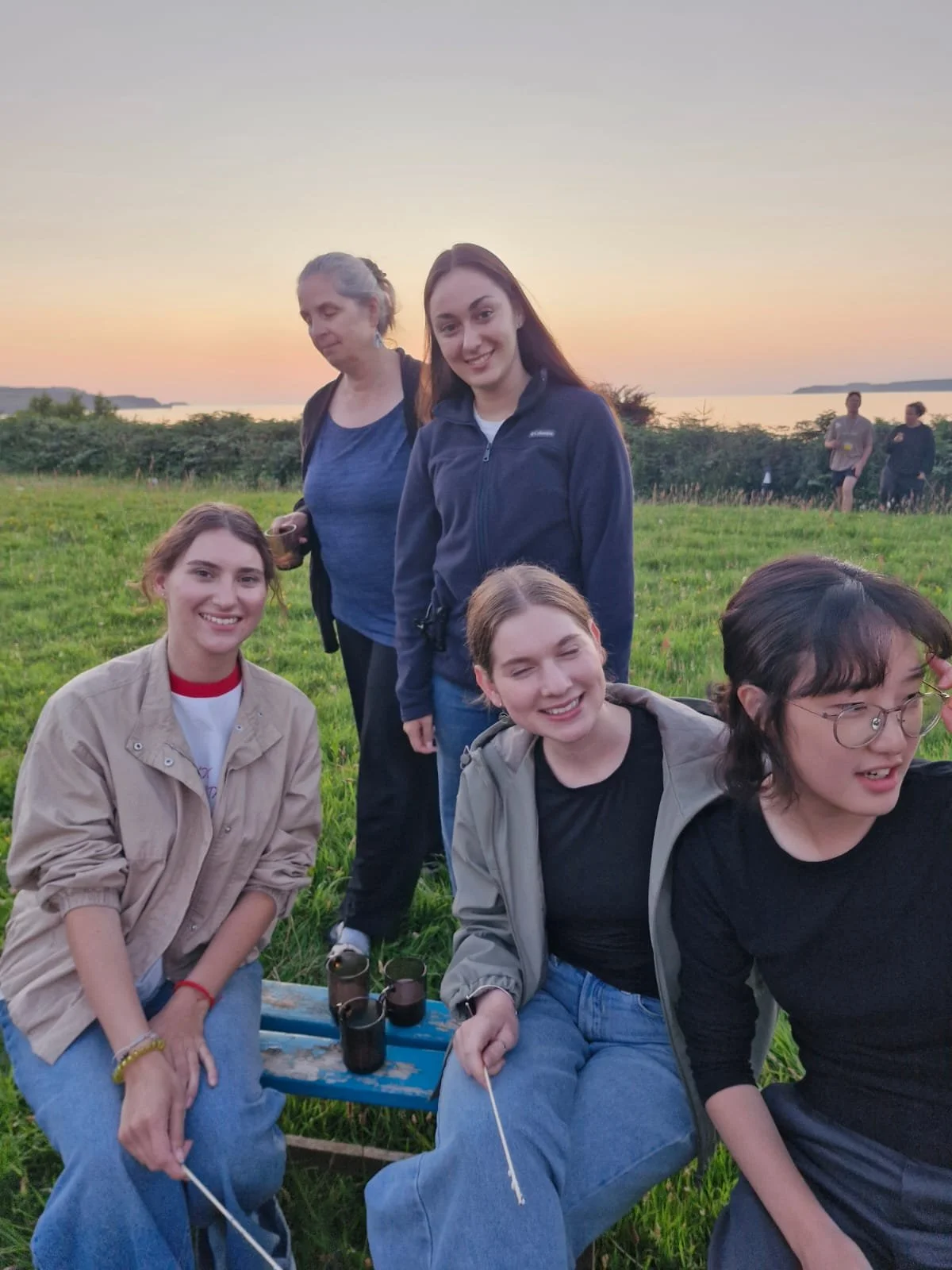 A group of five young women and one older woman outdoors near a body of water at sunset. They are smiling, sitting and standing on grassy field, with three binoculars and a picnic table in front of them.