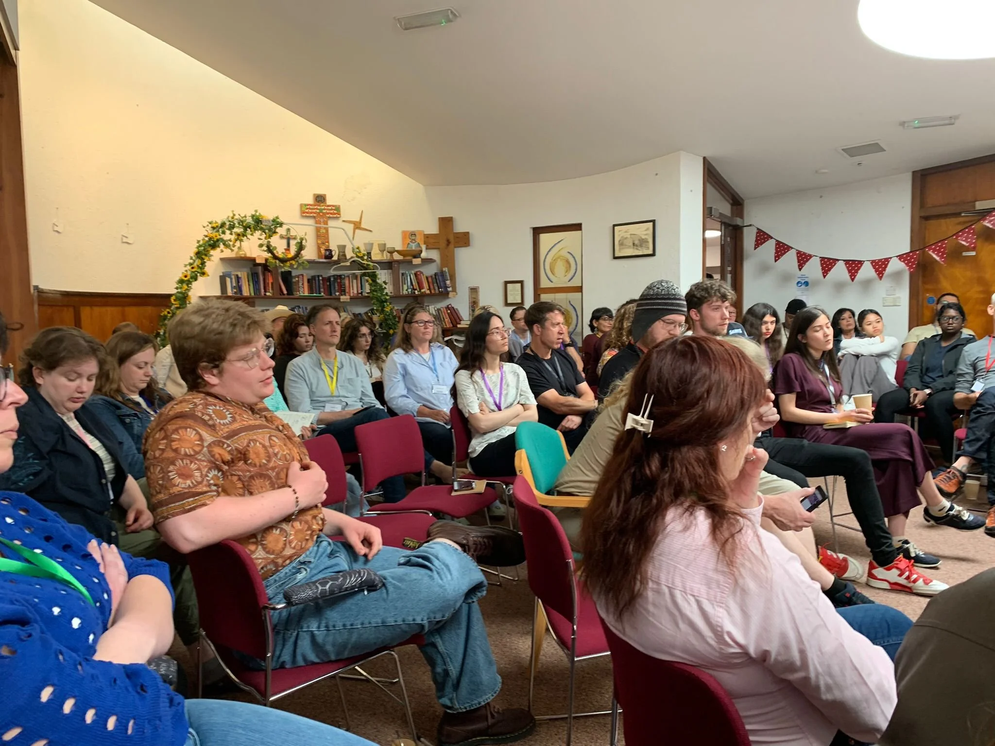 Group of people attending a religious or spiritual event in a room with church decor, bookshelves, and a wooden cross on the wall.