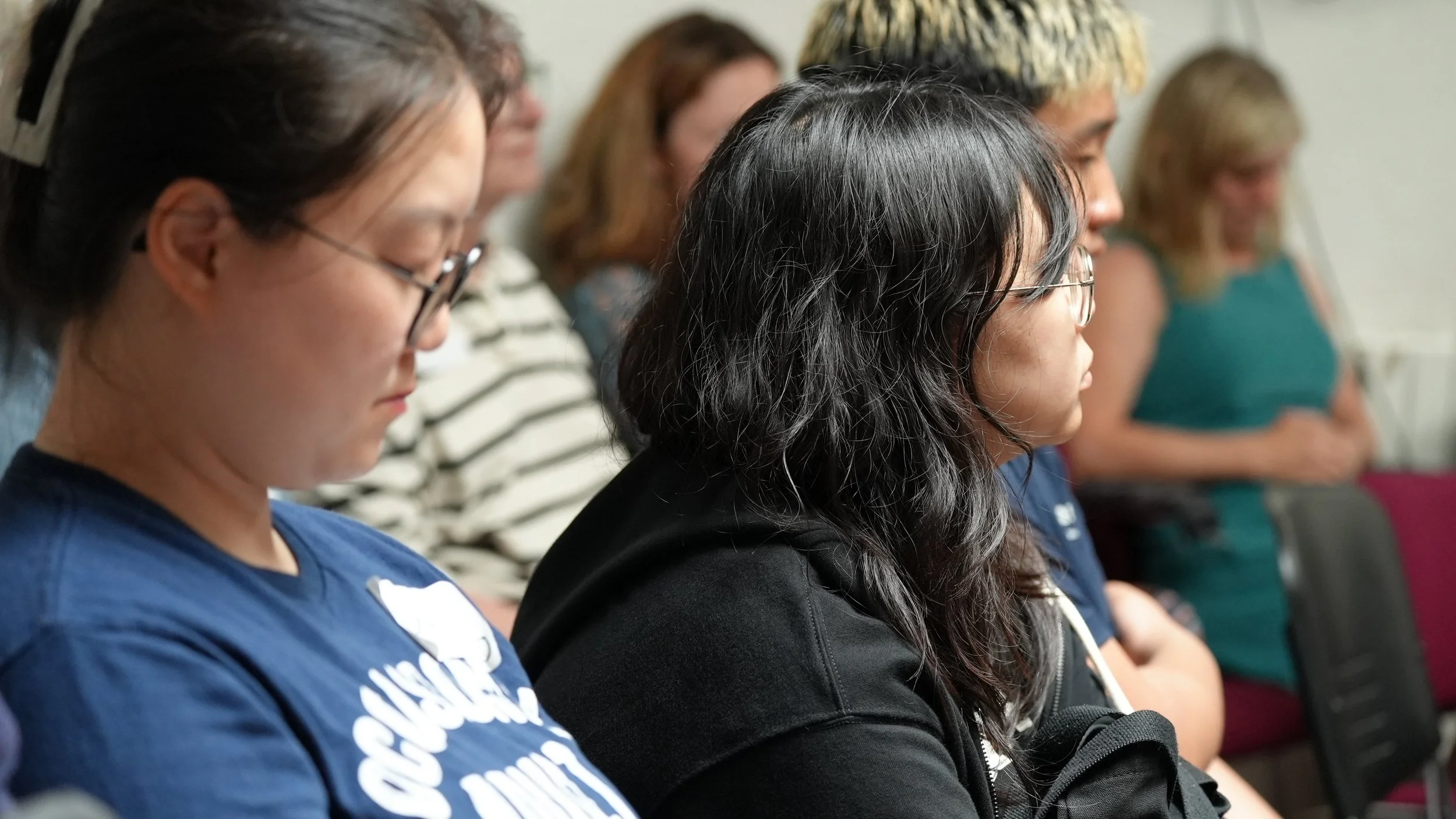 A group of people sitting in a row, with their eyes closed, attending a meditation or mindfulness session.